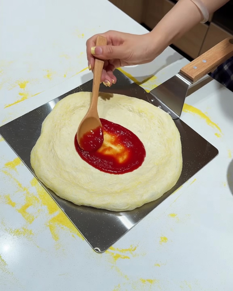 Une cuillère en bois étalant la sauce tomate rouge sur une pâte étirée.