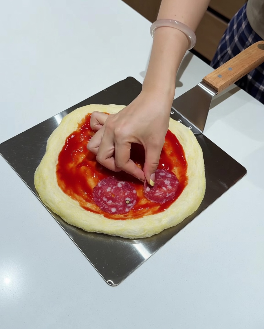 Une main plaçant des tranches de salami sur la base de pizza garnie de sauce.