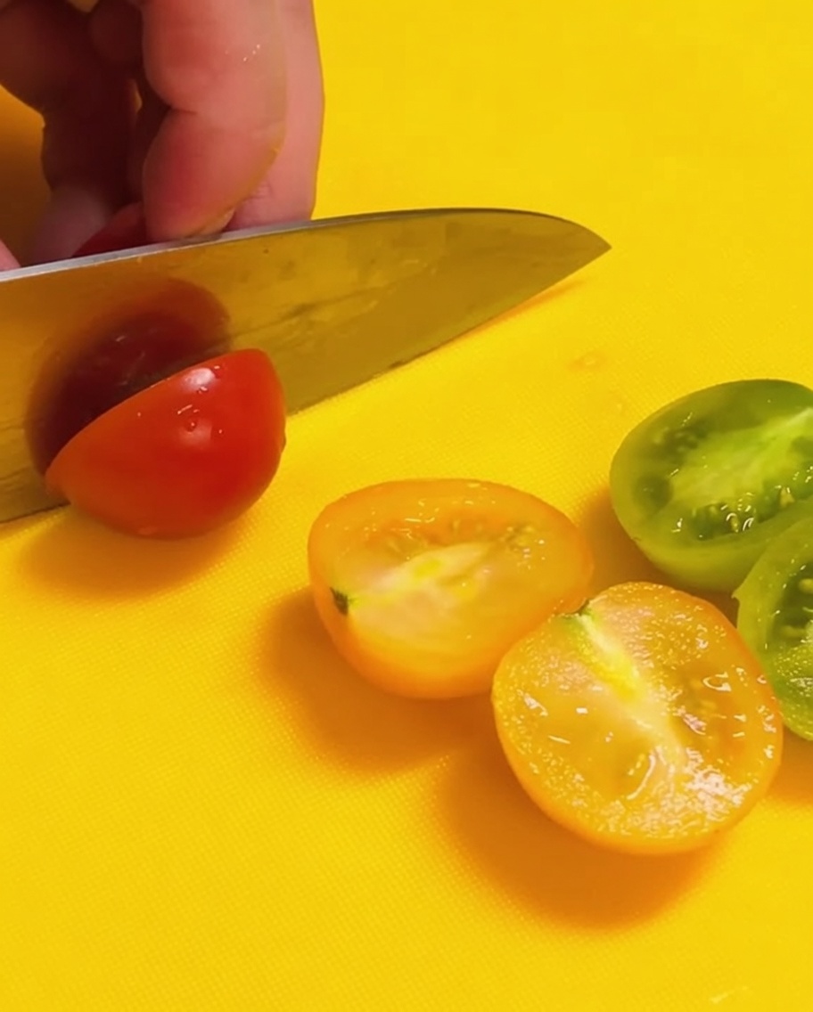 Un couteau coupant des tomates cerises rouges et vertes en deux sur une planche à découper jaune vif.