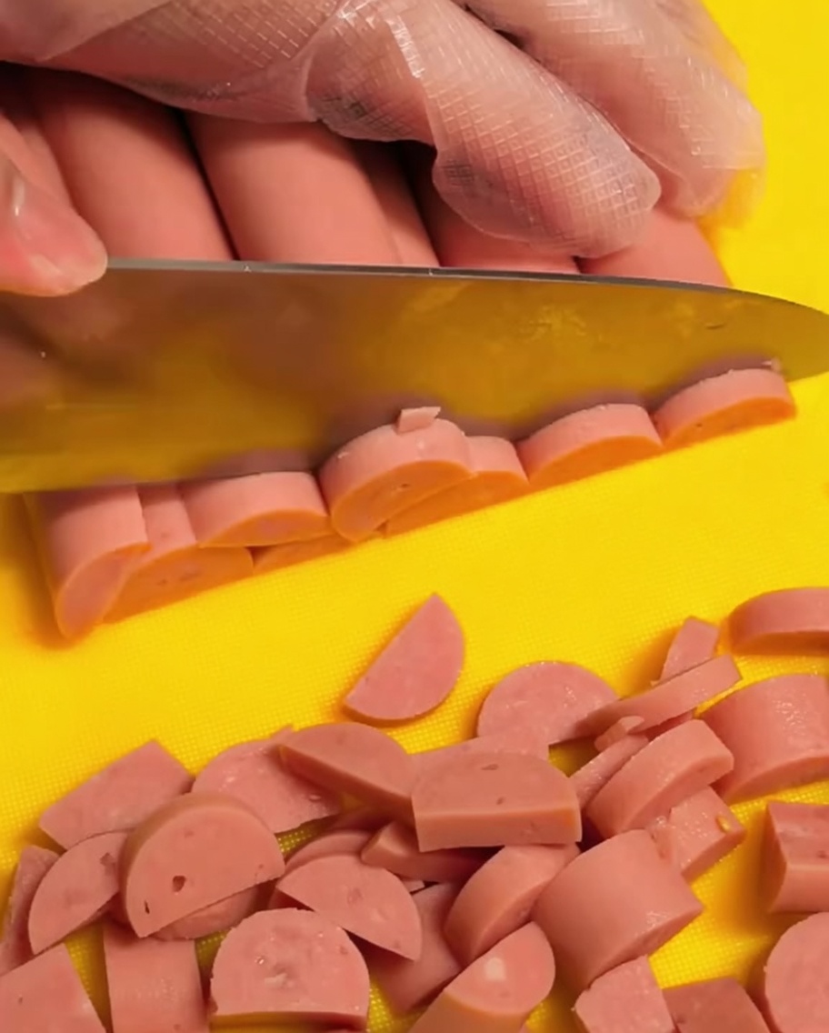 Une main portant un gant transparent découpant des saucisses de jambon roses en petites demi-lunes sur une planche à découper jaune.