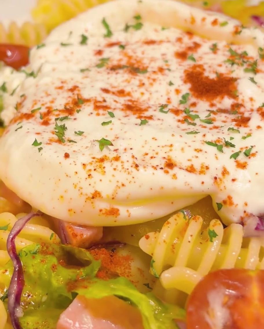 Gros plan du plat de pâtes froides terminé avec un cœur de burrata crémeux, garni de persil et de piment rouge, avec un dernier filet doré dhuile dolive.