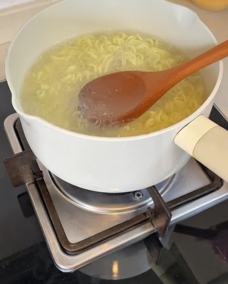 Une louche en bois versant de leau de cuisson des nouilles, chaude et trouble, dans le bol contenant la base.
