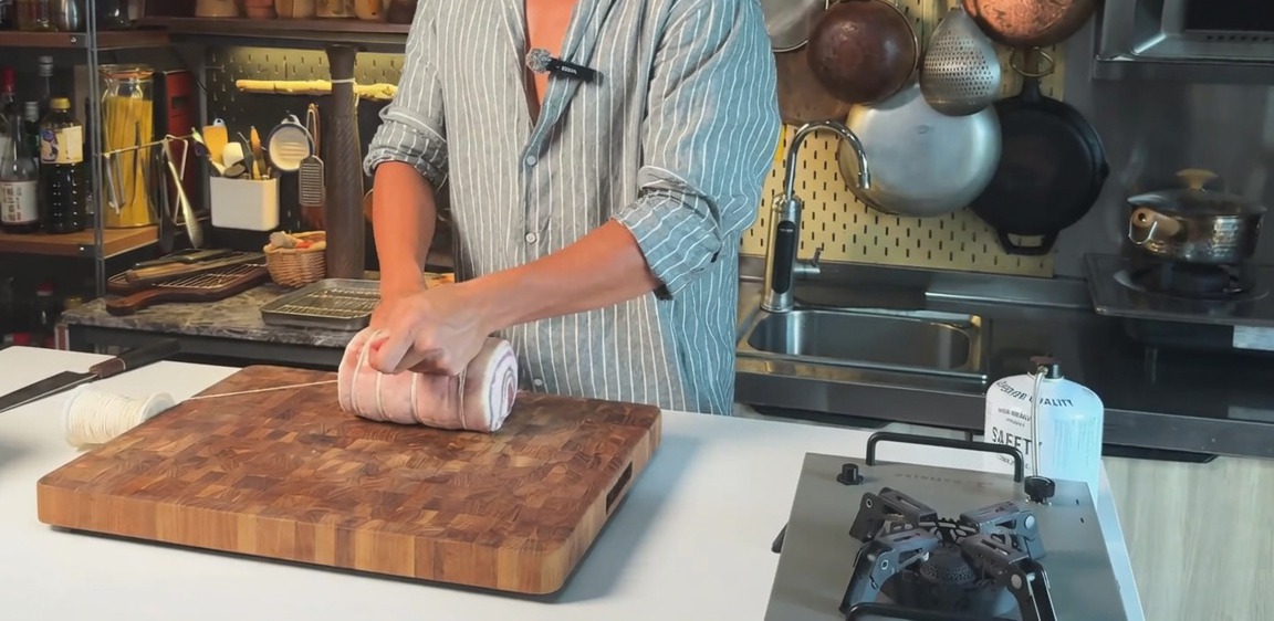 Une poitrine de porc crue roulée et ficelée horizontalement avec de la ficelle de cuisine sur une planche à découper en bois.