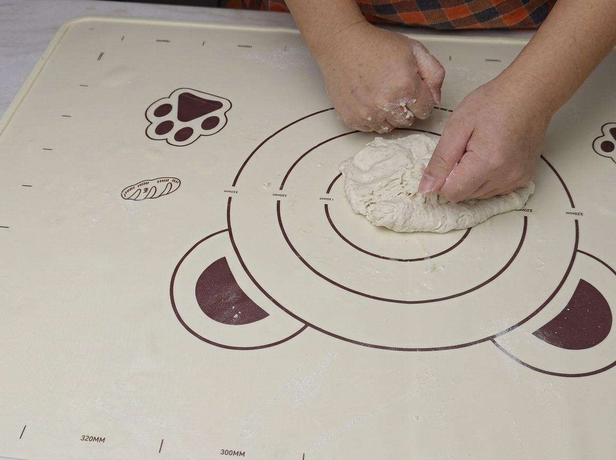 Deux mains pressant et pliant fermement une boule de pâte rugueuse sur un tapis en silicone.