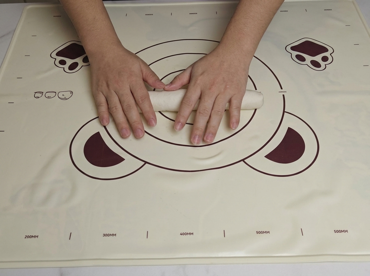 Deux mains roulant une pâte lisse en un long cylindre épais sur un tapis de cuisson.