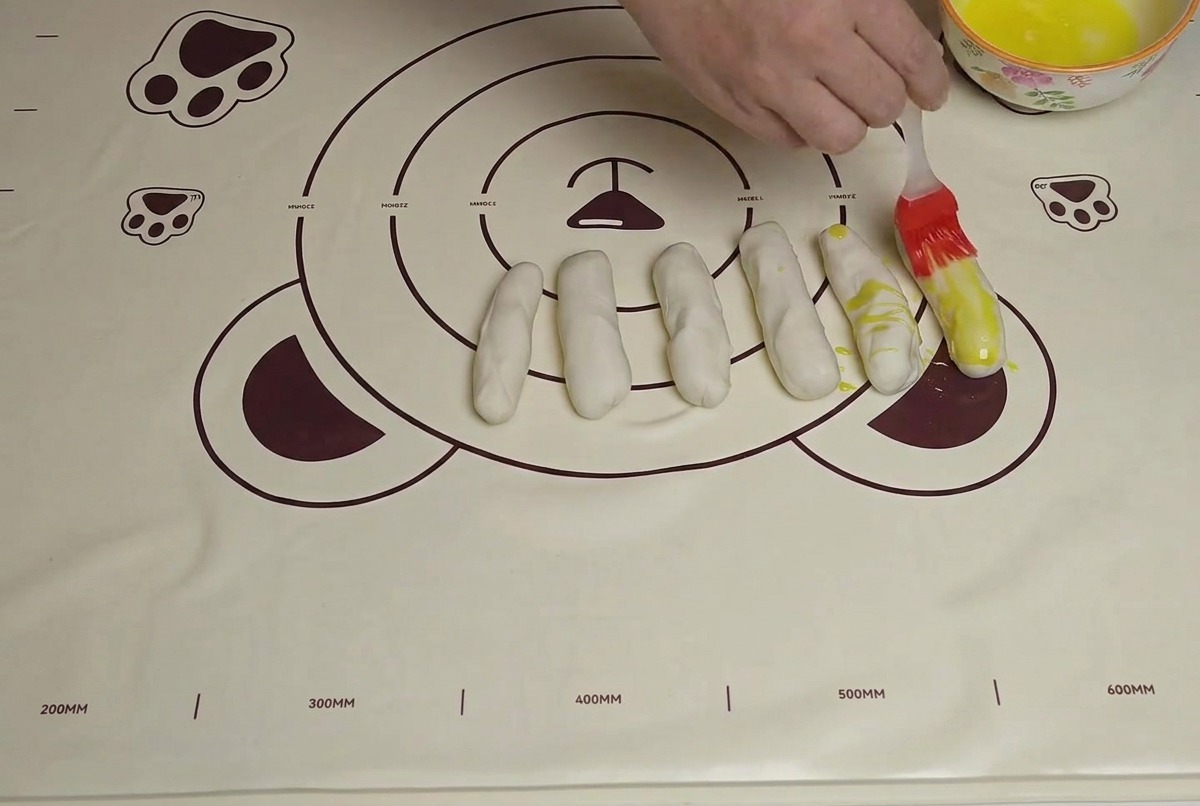 Plusieurs bandes de pâte disposées sur un tapis, badigeonnées dhuile à laide dun pinceau en silicone.