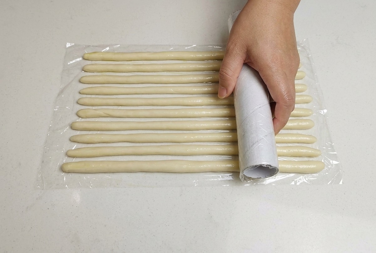 Une main plaçant un film plastique sur des cordes de pâte fines disposées sur une surface blanche.