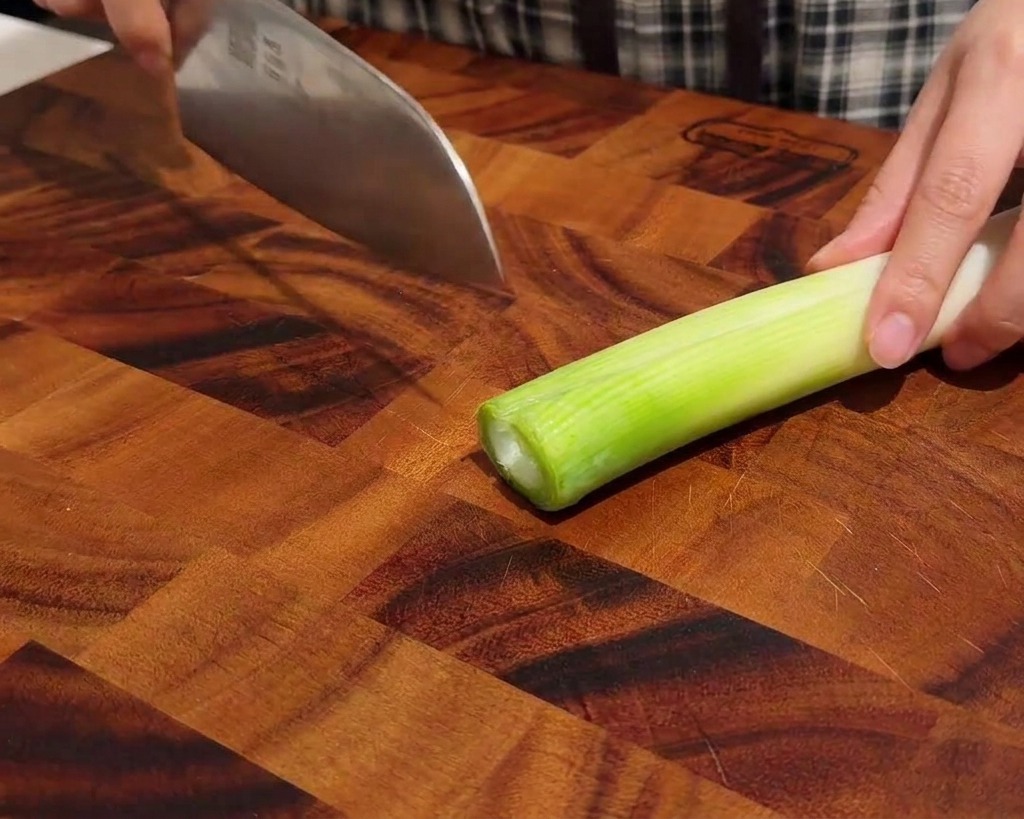 Mains coupant soigneusement un oignon vert en fines rondelles sur une planche à découper en bois foncé.
