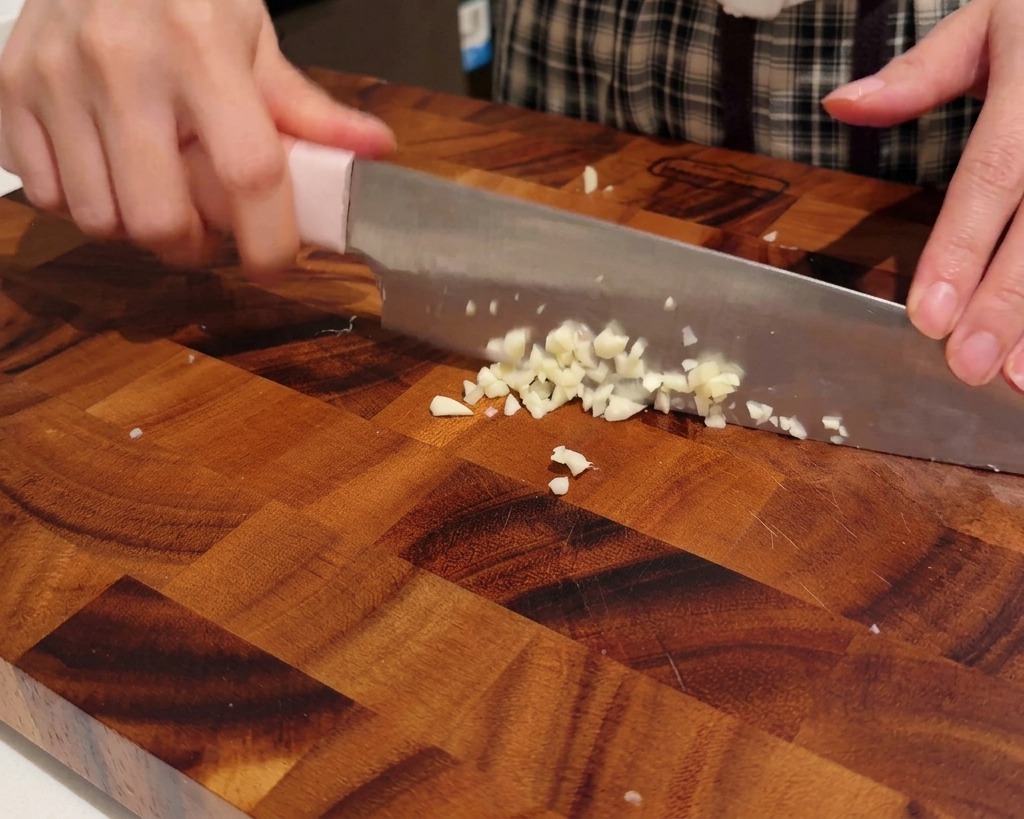 Mains tenant un couteau, hachant des gousses dail en petits morceaux fins sur une planche à découper en bois.
