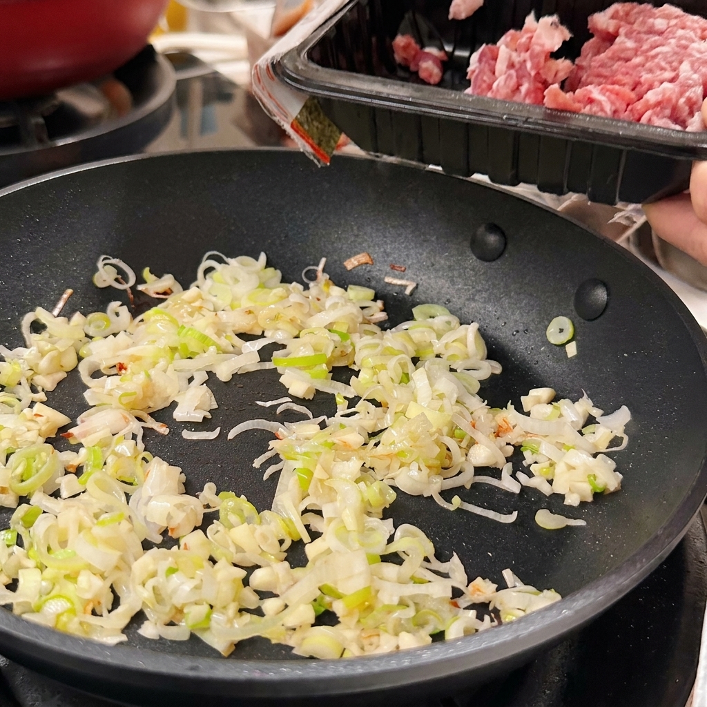 Porc haché cru versé dune barquette en plastique noir dans une poêle contenant les oignons verts et lail sautés.