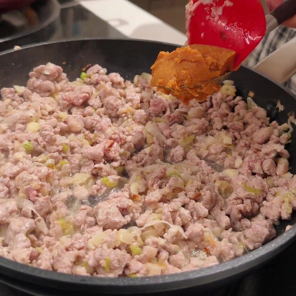 Une spatule rouge tenant une cuillerée de pâte miso au-dessus dune poêle remplie de porc haché doré.