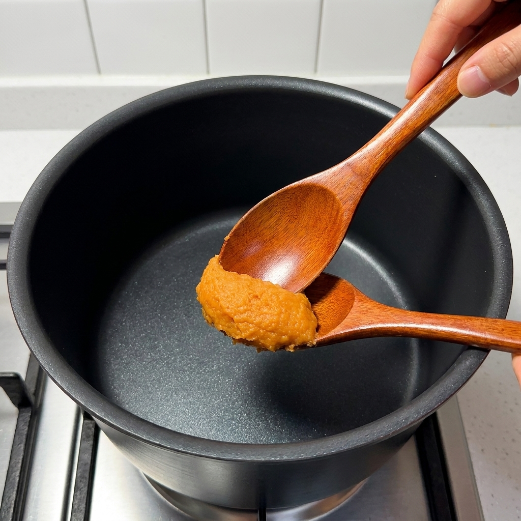 Une cuillère en bois remuant de la pâte miso, du mirin, du sucre, du bouillon dos de porc et du jus de yuzu dans une casserole sur une cuisinière.