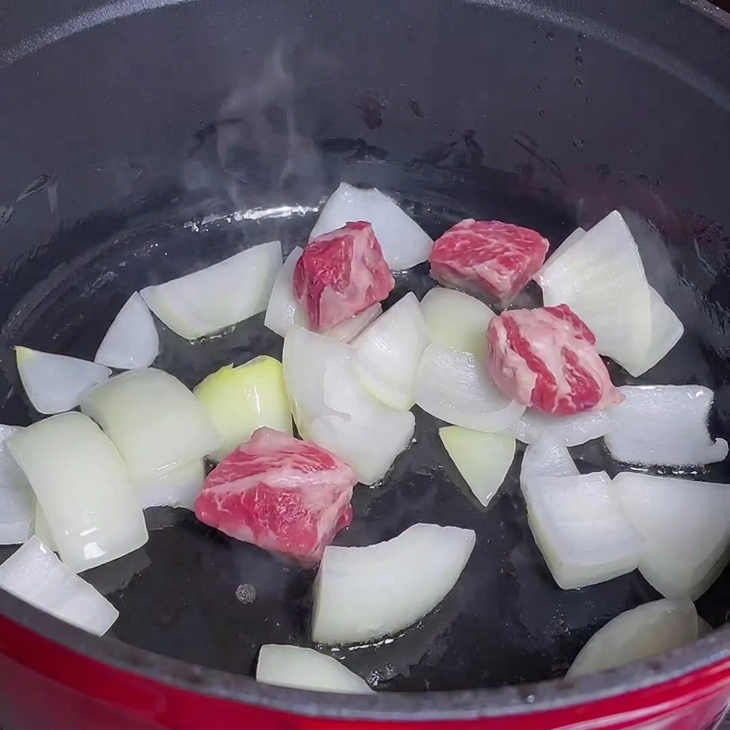 Cubes de bœuf cuisant avec des oignons dans une casserole rouge.