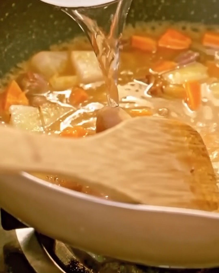 De leau versée dans une poêle remplie de bœuf, de légumes et de curry fondant, avec une spatule en bois visible.