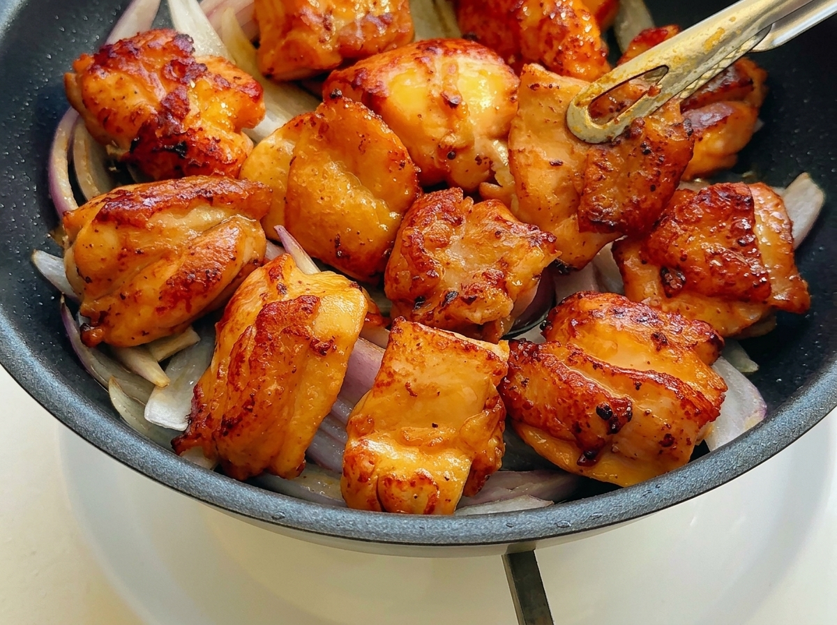 Morceaux de poulet poêlés disposés sur les oignons rouges sautés dans une poêle.