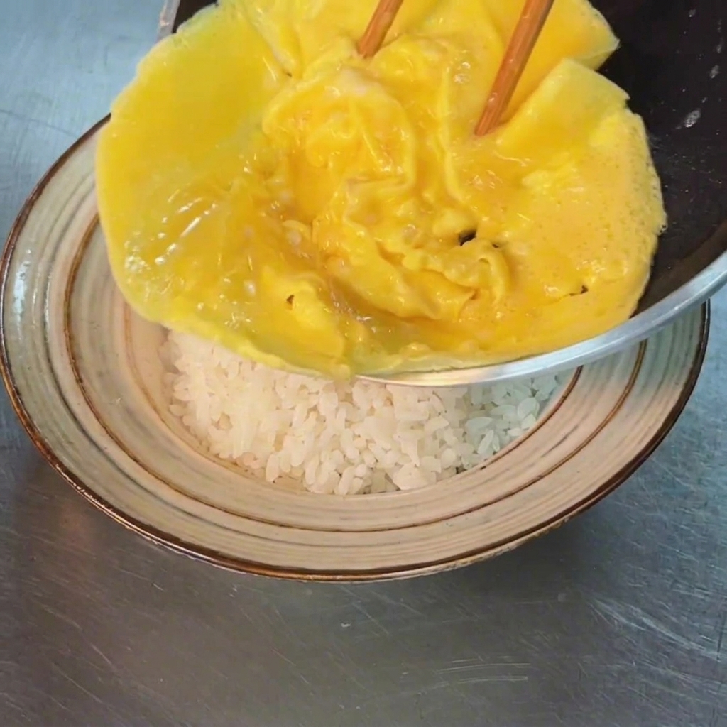 Lomelette tendre et baveuse glissée délicatement depuis la poêle sur un monticule de riz blanc dans un bol.