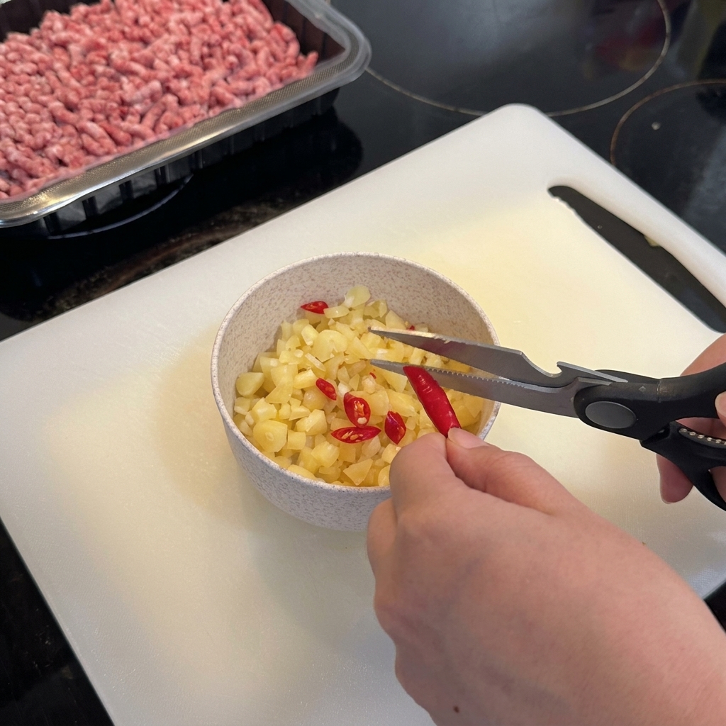 Ciseaux de cuisine coupant des piments oiseaux rouges dans un bol avec de lail haché.