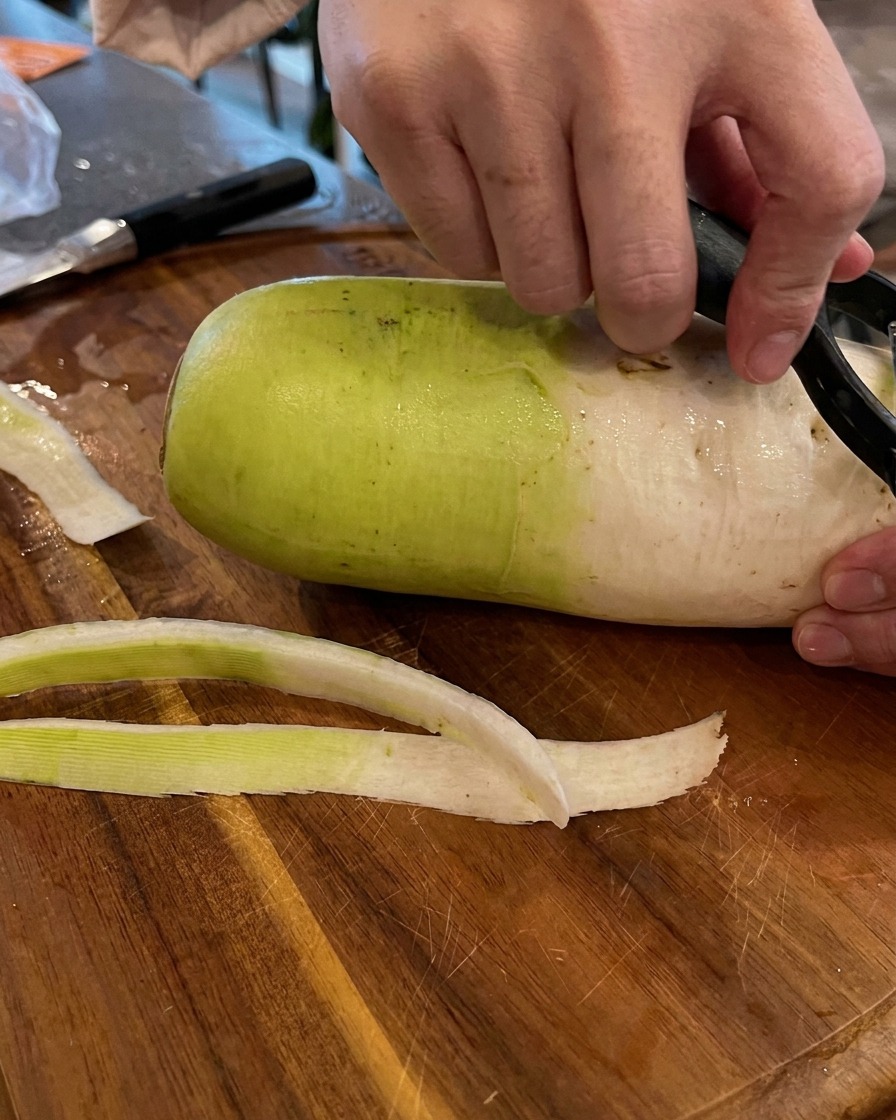 Mains utilisant un éplucheur à légumes noir pour retirer la peau dun gros radis sur une planche à découper en bois.