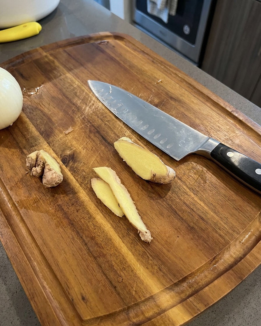 Fines tranches de gingembre frais reposant sur une planche à découper en bois à côté dun couteau de chef et dun demi-oignon.