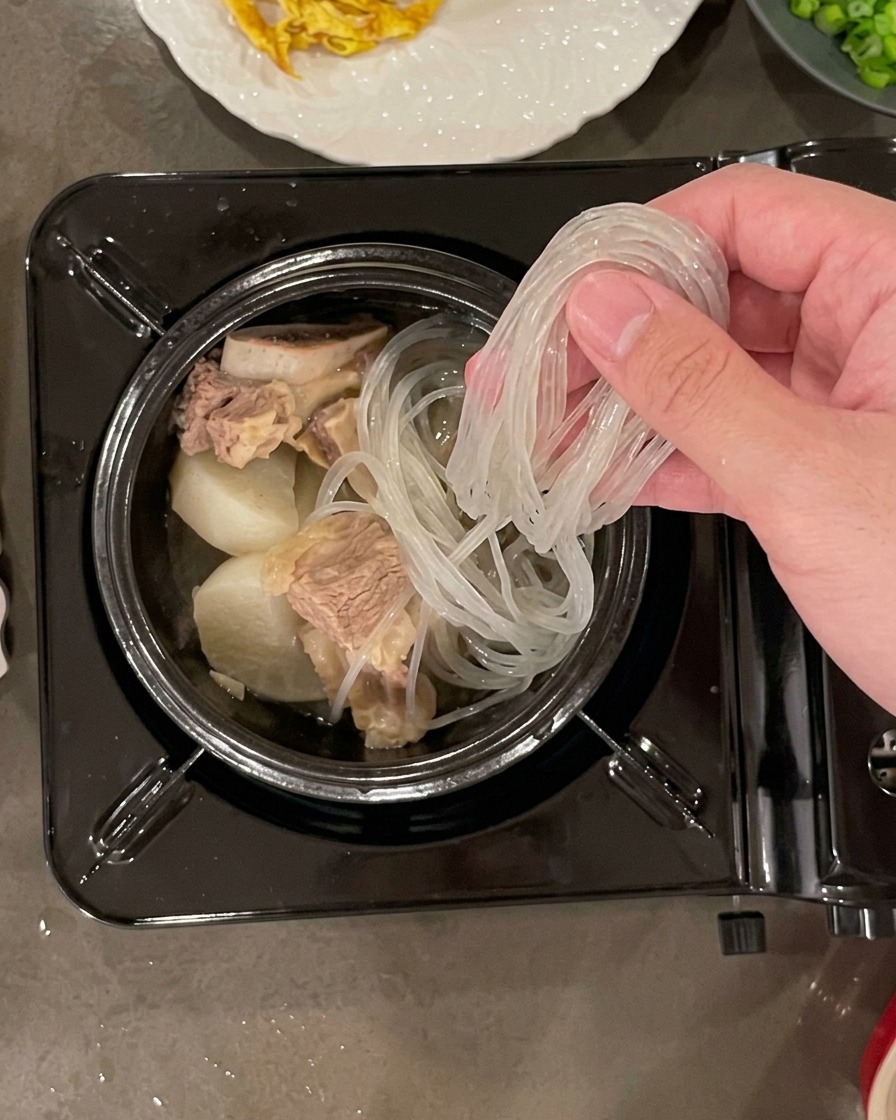 Main plaçant une portion de nouilles de verre trempées et translucides dans un pot en pierre rempli de bœuf et de morceaux de radis.