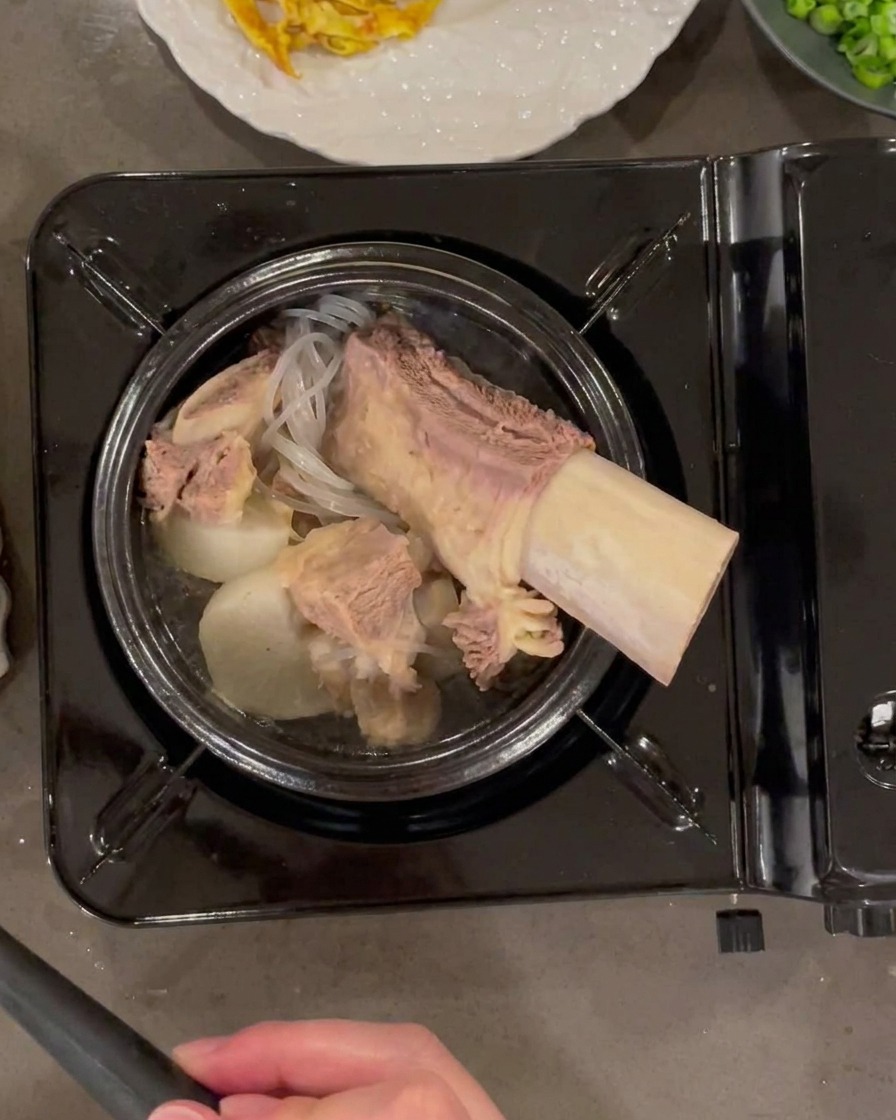 Louche versant un bouillon de bœuf clair et chaud dans un pot en pierre rempli de bœuf, de radis et de nouilles.
