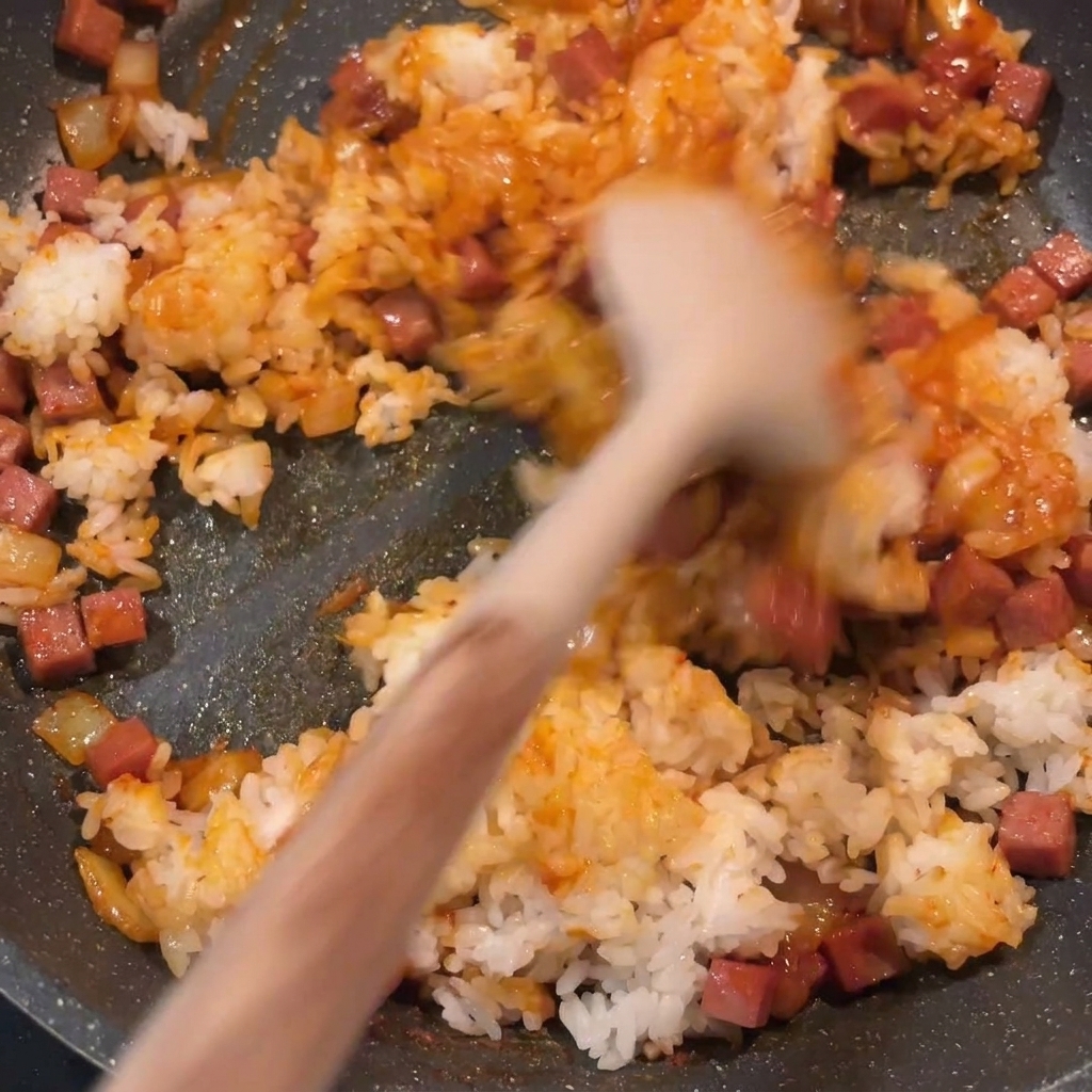 Le riz sauté au kimchi terminé en train dêtre mélangé dans une poêle, montrant une couleur rouge uniforme et des morceaux de spam et doignon bien répartis.