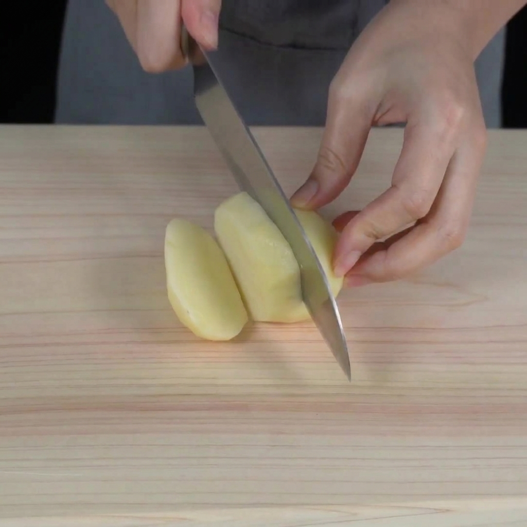 Mains utilisant un couteau de chef pour couper une pomme de terre crue pelée en petits cubes sur une planche à découper en bois.