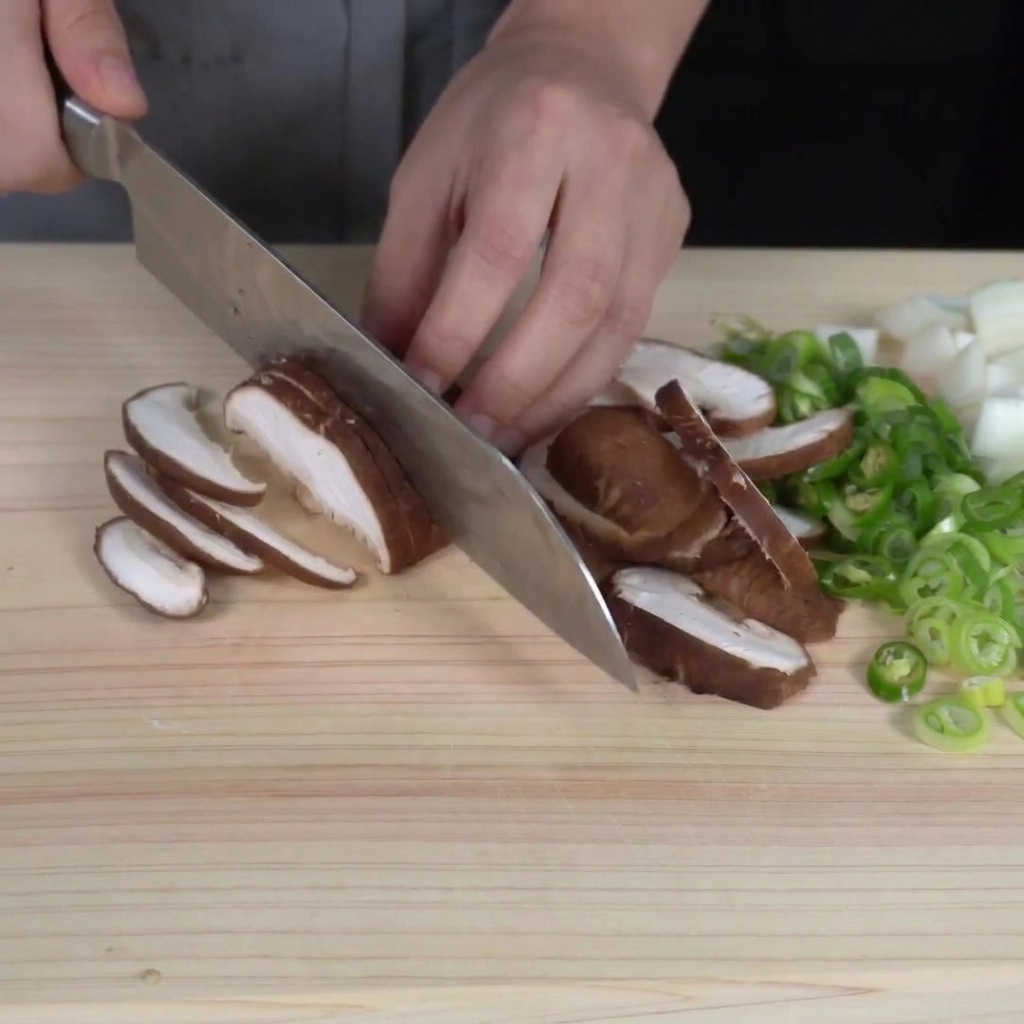 Tranchage fin de champignons shiitake frais sur une planche à découper, avec des légumes hachés visibles à proximité.