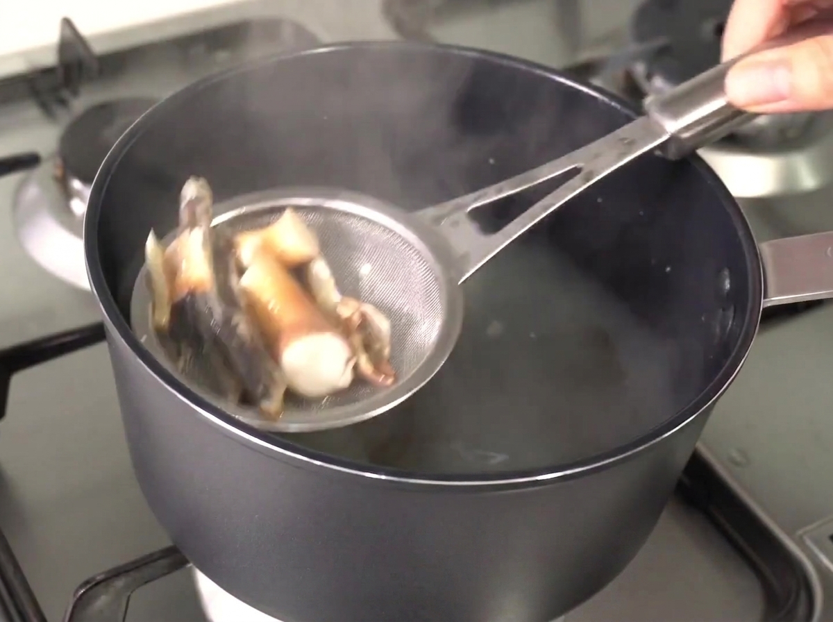 Une passoire en métal soulevant les anchois et le varech ramollis hors du bouillon chaud et clair.