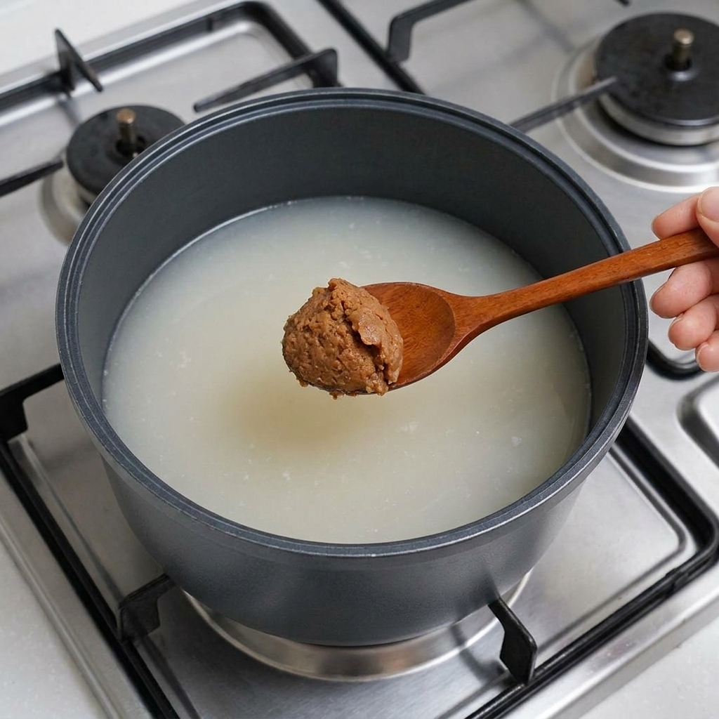 Une cuillère en bois tenant une généreuse dose de pâte de soja brune étant abaissée dans le bouillon.