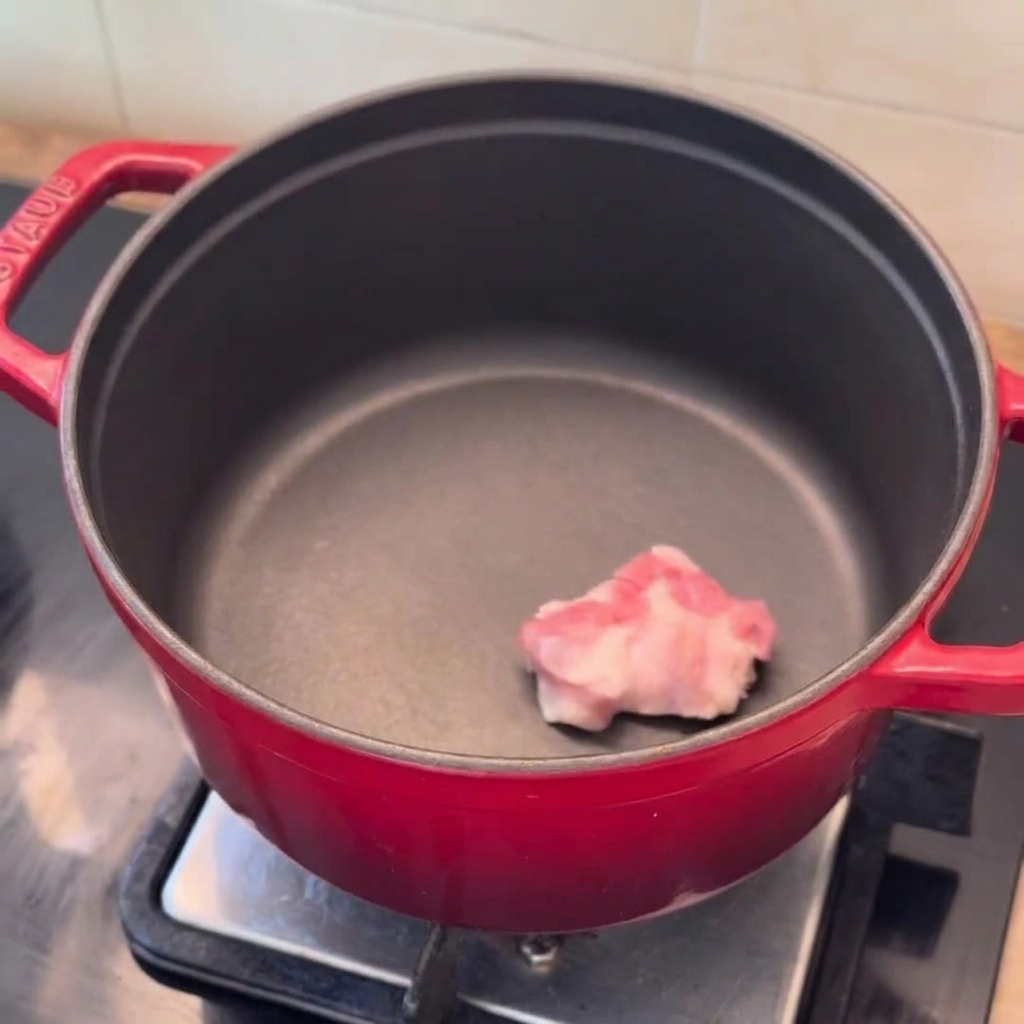 Des tranches de poitrine de porc, des quartiers doignon et de lail haché prêts dans une marmite rouge préchauffée.