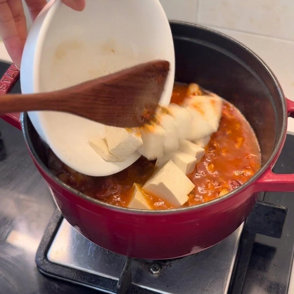 Des cubes de tofu frais sont délicatement versés dun bol blanc dans une marmite rouge profonde de ragoût de porc au kimchi bouillonnant.