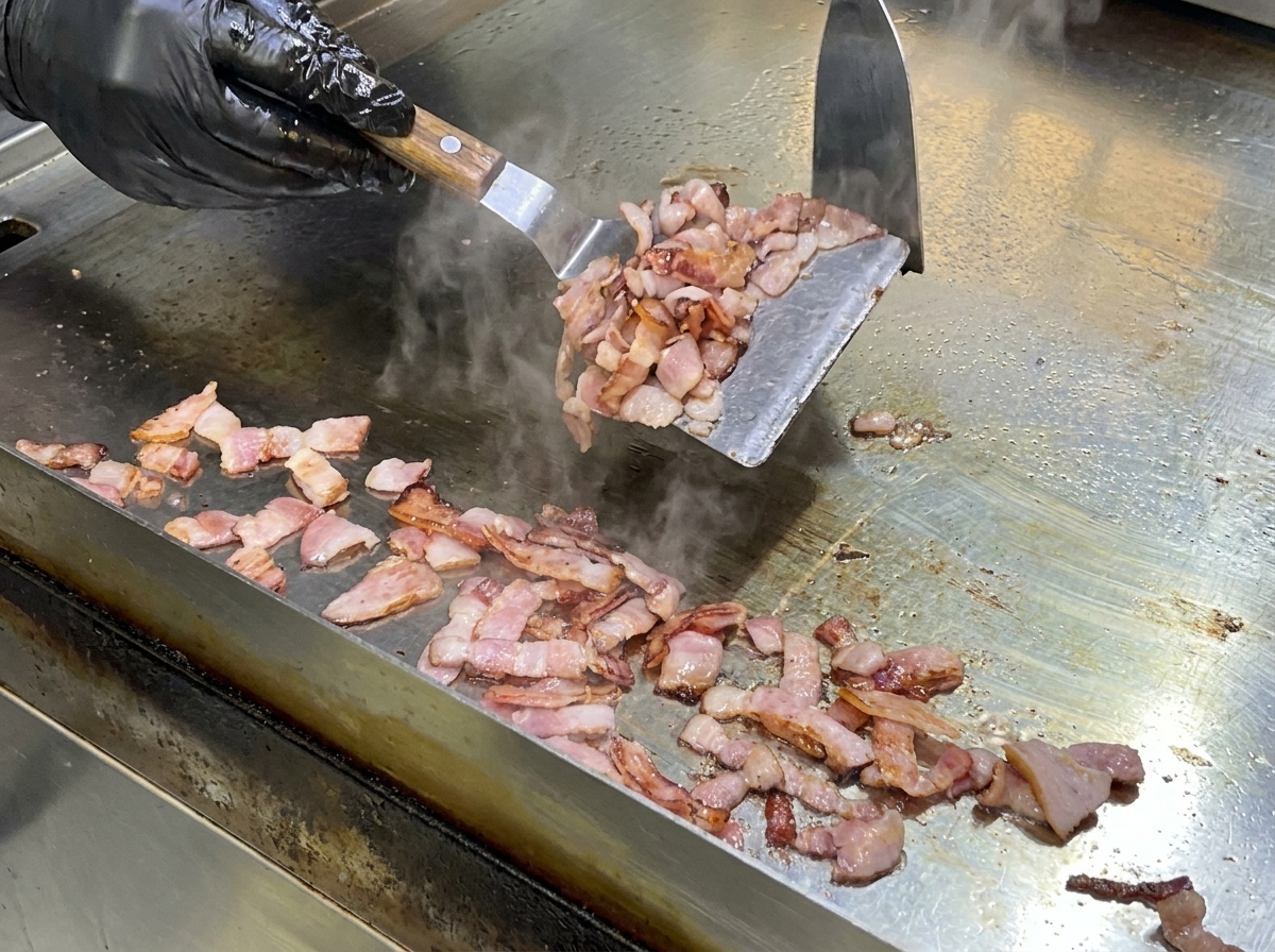 Une spatule métallique remuant les morceaux de bacon sur une plaque chaude.