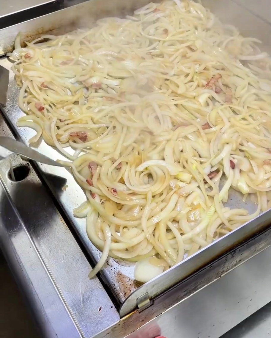 Une spatule remuant un monticule doignons blancs ramollis mélangés avec des morceaux de bacon sur une plaque en métal fumante.