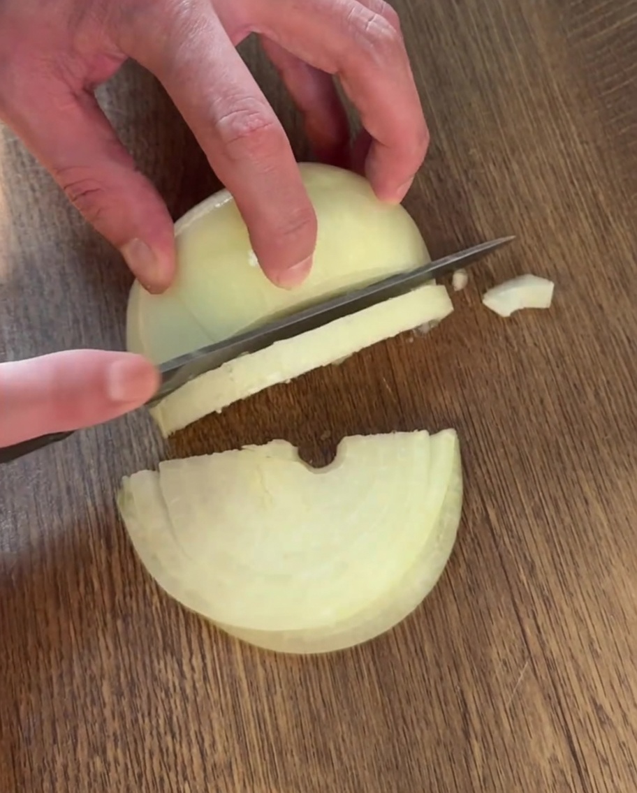 Une main utilisant un couteau pour trancher un oignon en fines lamelles sur une planche à découper en bois.