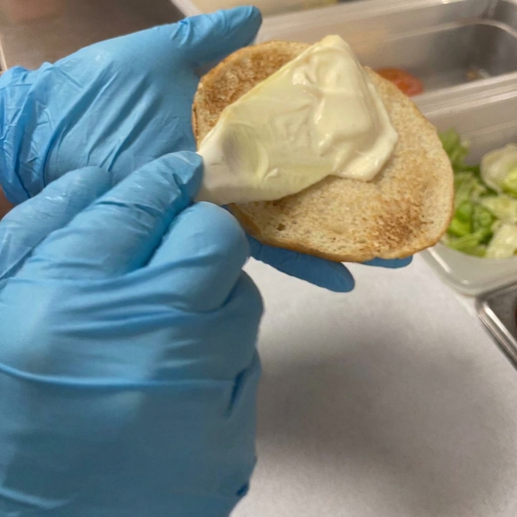 Une spatule en plastique étalant de la mayonnaise sur un pain à burger toasté.