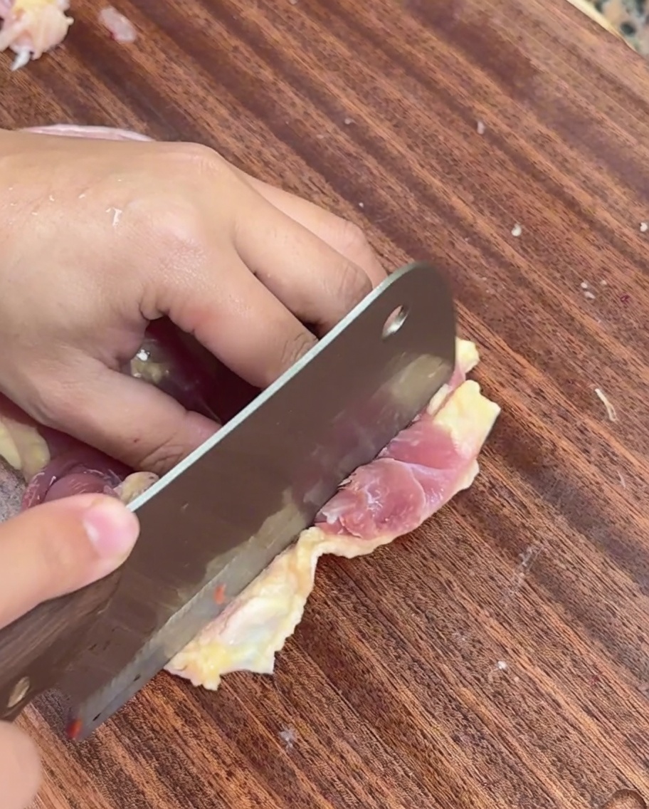 Une main utilisant un couperet pour trancher la chair de poulet désossée en fines lamelles sur une planche en bois.