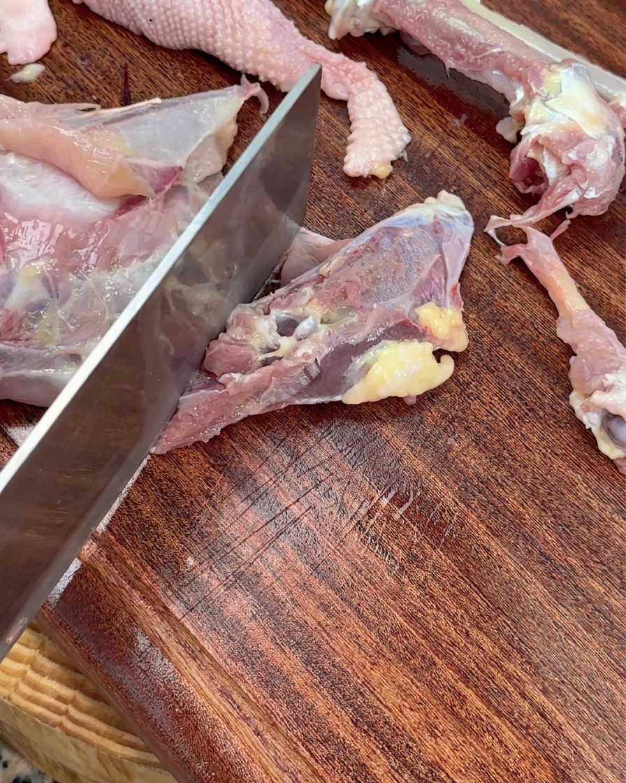 Un couperet hachant un squelette de poulet cru en gros morceaux sur une planche à découper en bois.