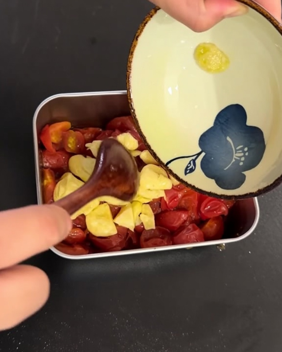 Lhuile dolive infusée à lail et lail émincé versés sur les tomates cerises cuites dans un plat en métal.