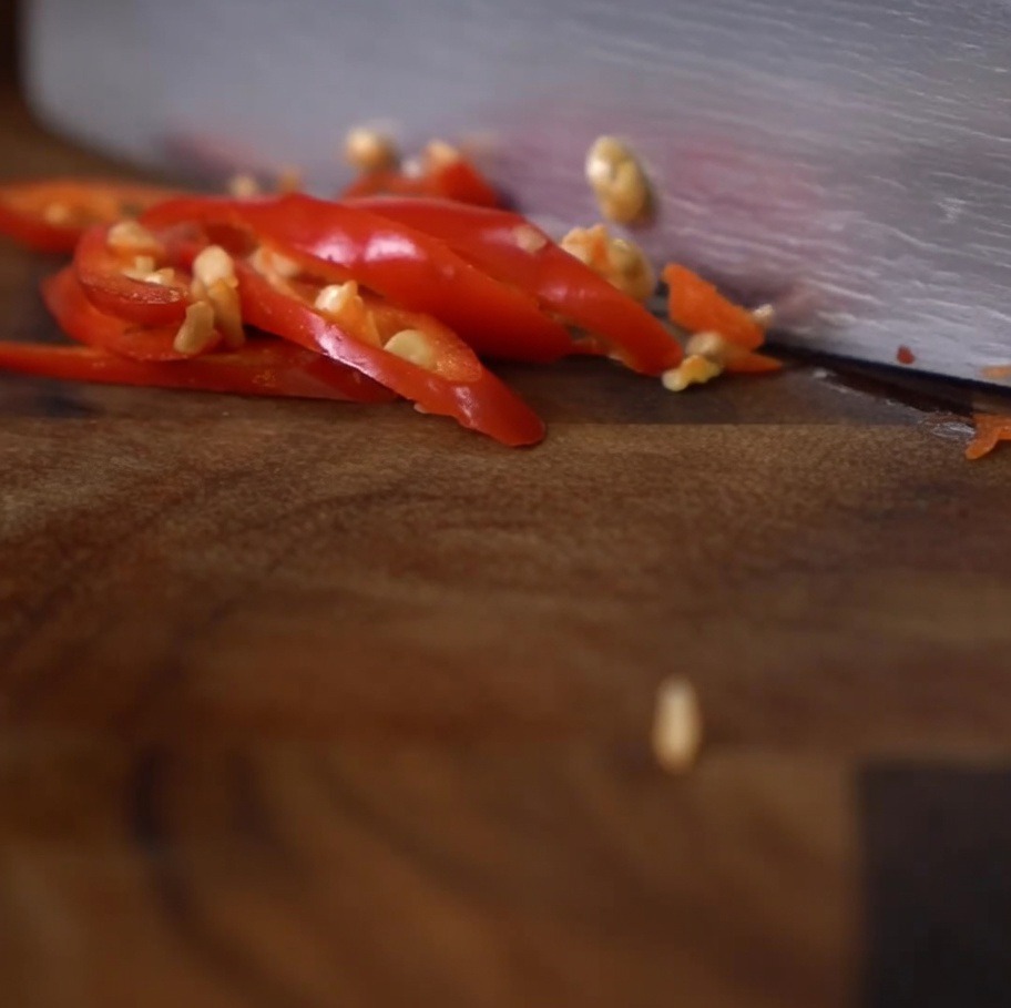 Gros plan dun couteau coupant des piments rouges vifs en petites rondelles sur une planche à découper en bois foncé.