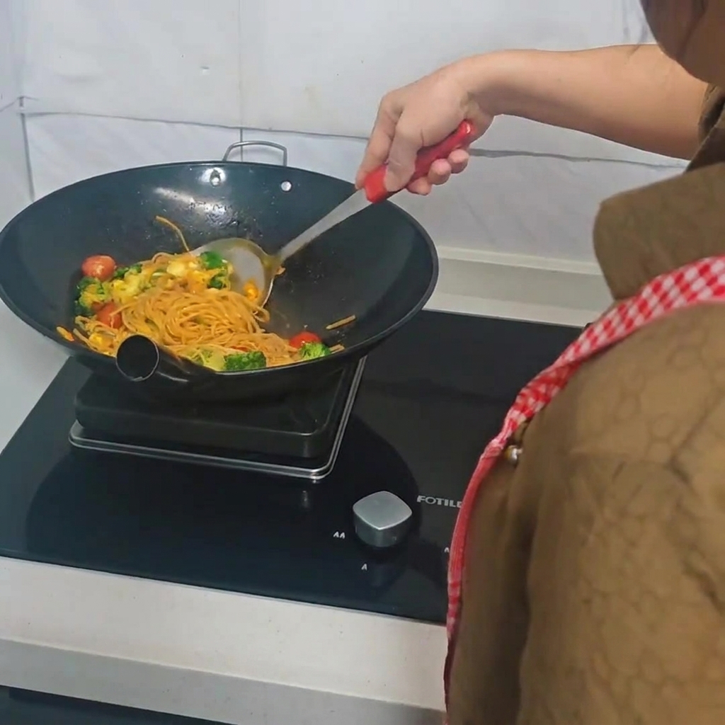 Une personne utilisant une spatule pour faire sauter des spaghettis, du brocoli, des tomates cerises et des œufs dans un wok.
