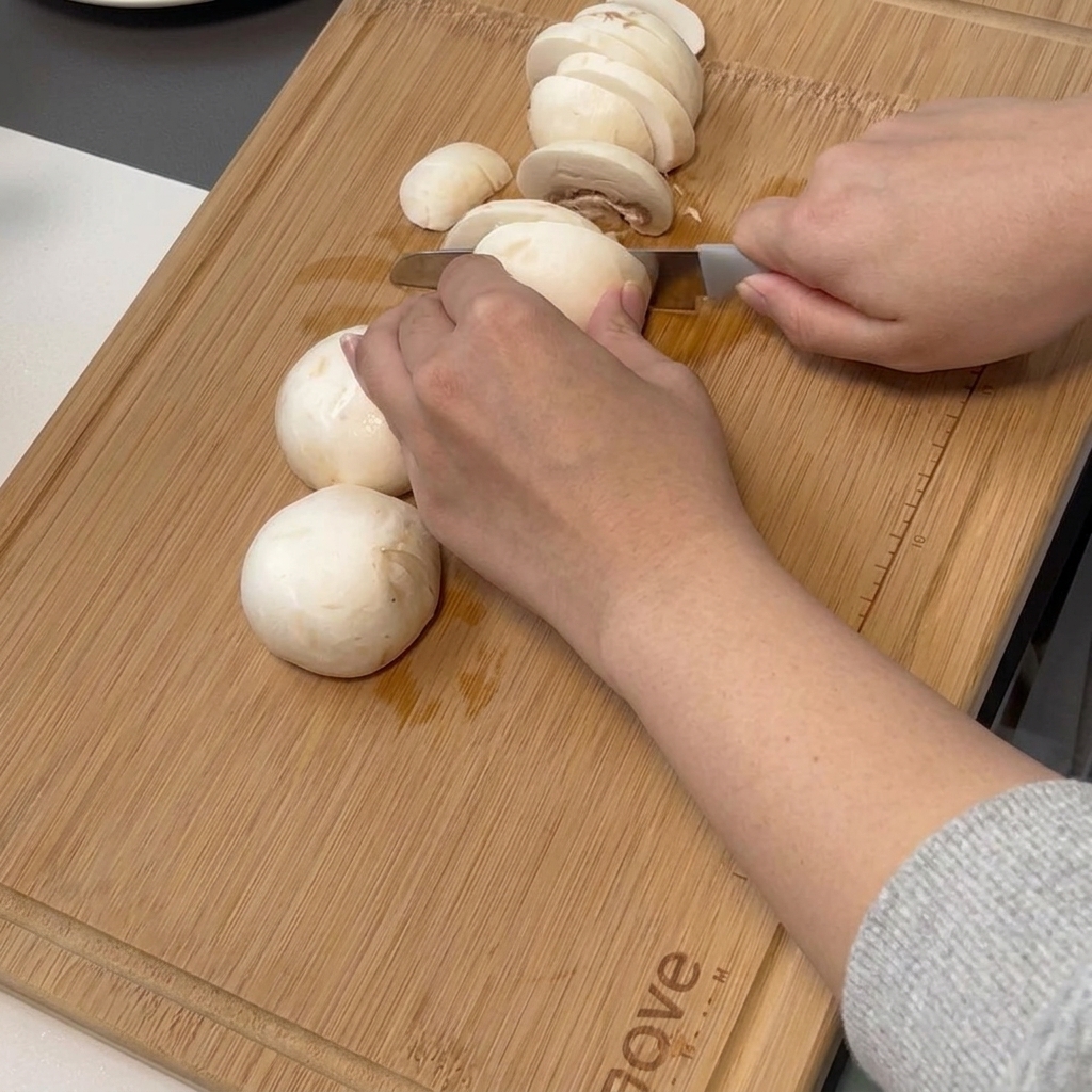 Une main utilisant un couteau de chef pour trancher finement des champignons de Paris blancs sur une planche à découper en bambou.