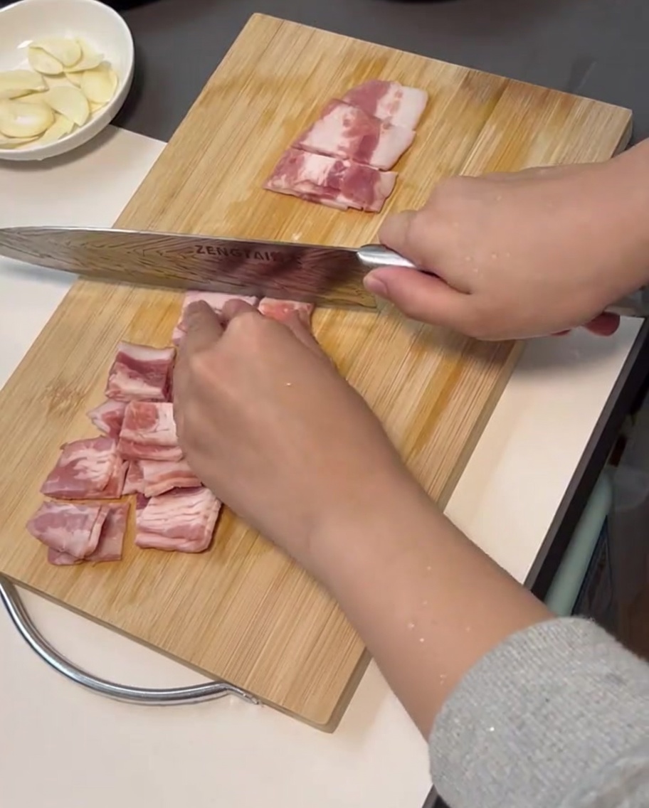 Des tranches de bacon frais coupées en petits morceaux réguliers sur une planche à découper avec un couteau.