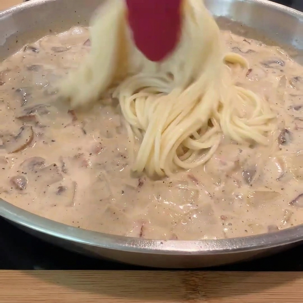 Des spaghettis cuits incorporés dans la poêle remplie de sauce crémeuse et de garniture.