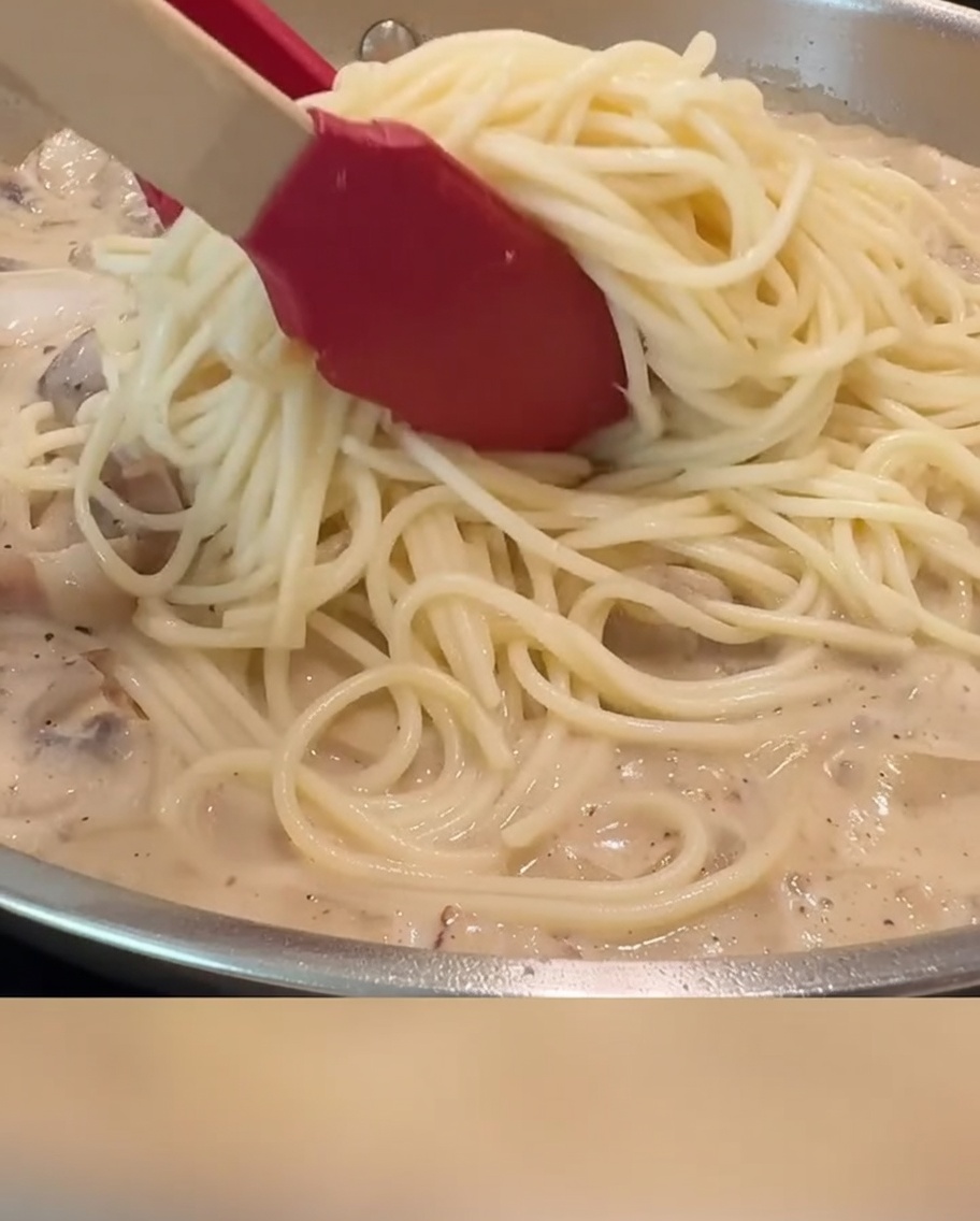 Des spaghettis enrobés de sauce crémeuse, de lardons et de champignons, mélangés dans une poêle à laide de pinces rouges.