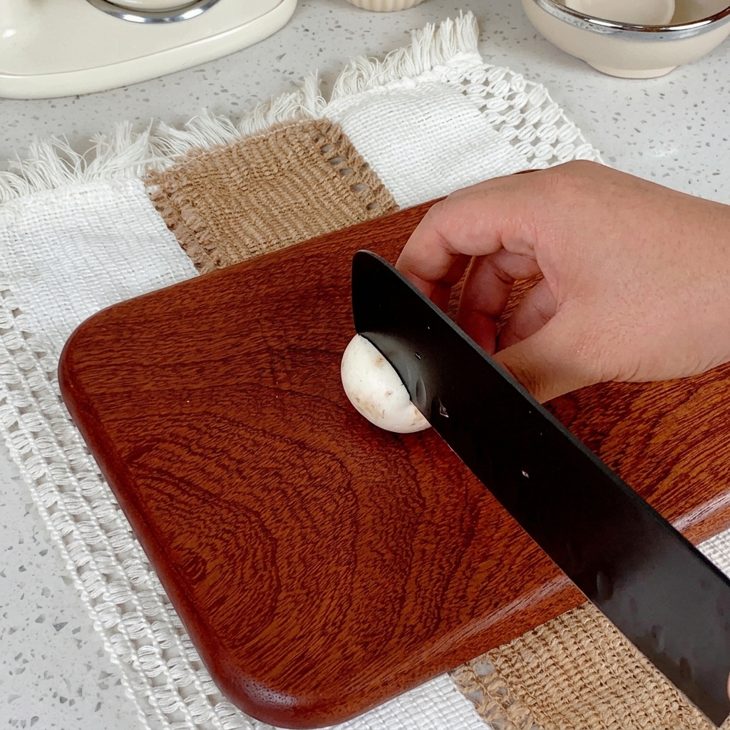 Une main utilisant un couteau noir pour trancher un champignon de Paris sur une planche en bois.