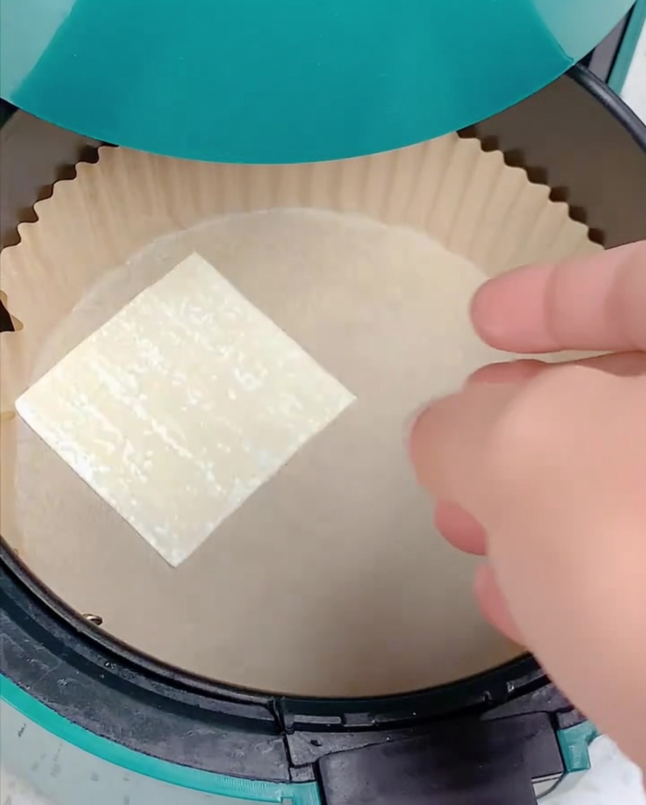 Une main plaçant une feuille de wonton carrée dans un panier de friteuse à air recouvert de papier sulfurisé.