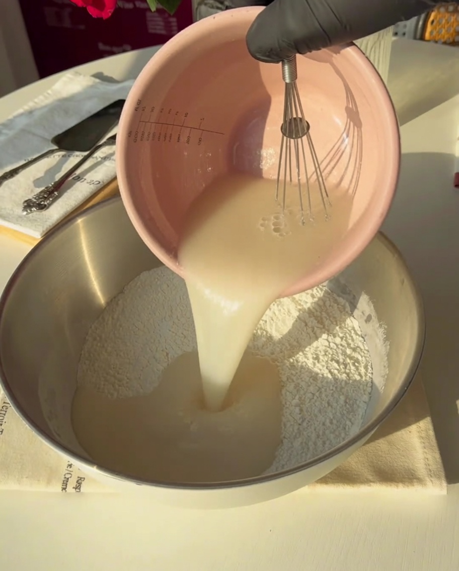 Une main gantée versant le mélange levure-eau dans un grand bol en métal contenant de la farine.