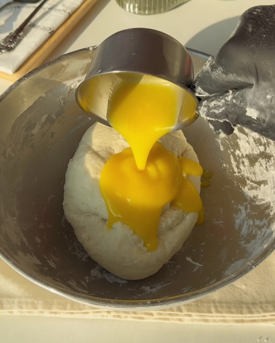 Une main gantée versant du beurre fondu doré sur la pâte.