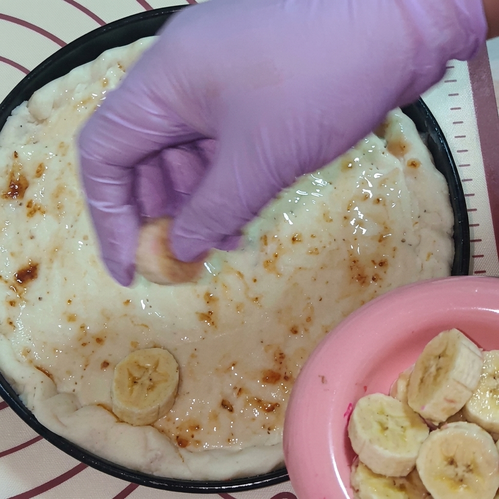 Une main portant un gant violet disposant des tranches de fruits frais, dont de la banane et du fruit du dragon, sur la pâte à pizza couverte de confiture.